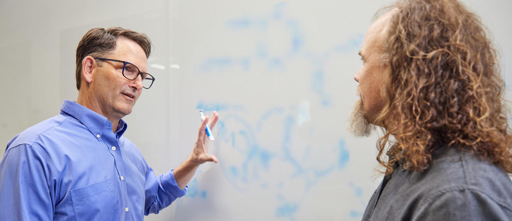 澳门正规博彩十大排行平台 scientists Brett Crawford and Dan Wendt talking by a whiteboard with a scientific formula